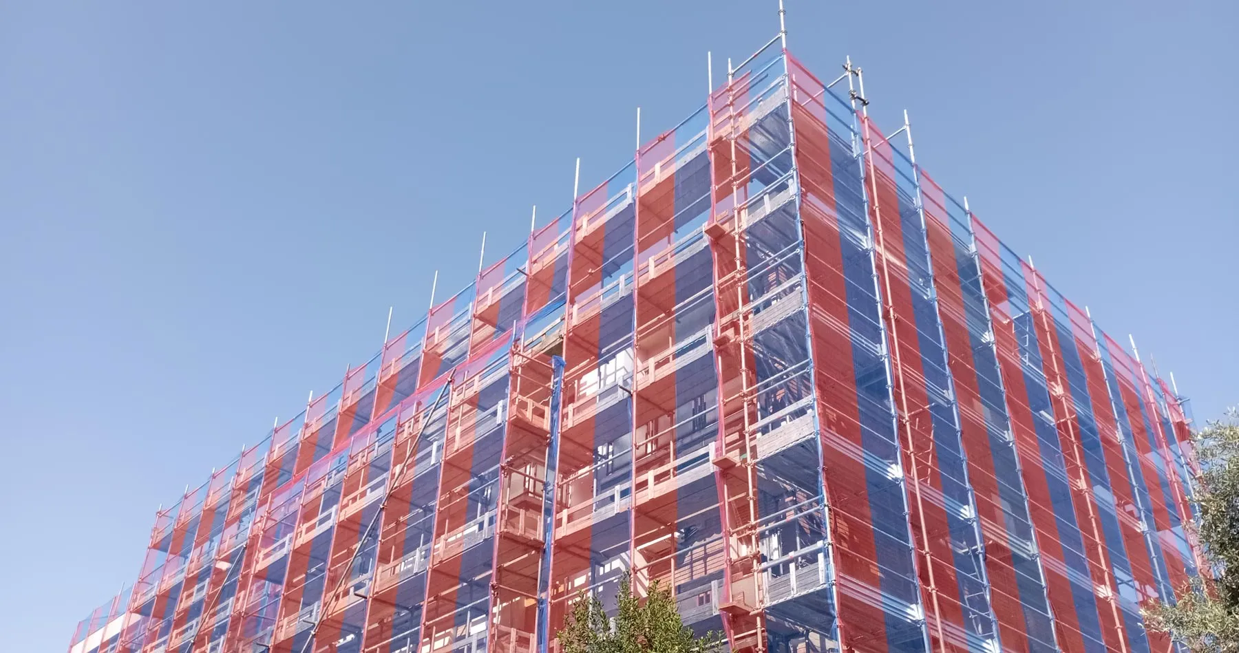 Edificio fasciato dal ponteggio con il telo a strisce blu e rosse, visto dal basso
